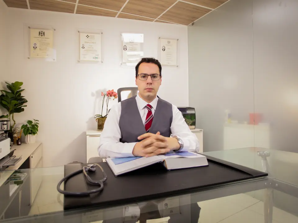 Oncólogo en su consultorio privado, sentado frente al escritorio con un libro abierto y un estetoscopio, como parte de una campaña de publicidad para médicos especialistas.